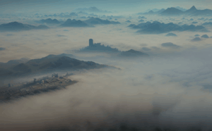 A smoggy view from several thousand feet. Through the smog we can see a town on the left hand side and in the centre is a hill also visible through the smog. A tower or grain silo is visible on the hill. Other hills are poking through the smog in the distance.